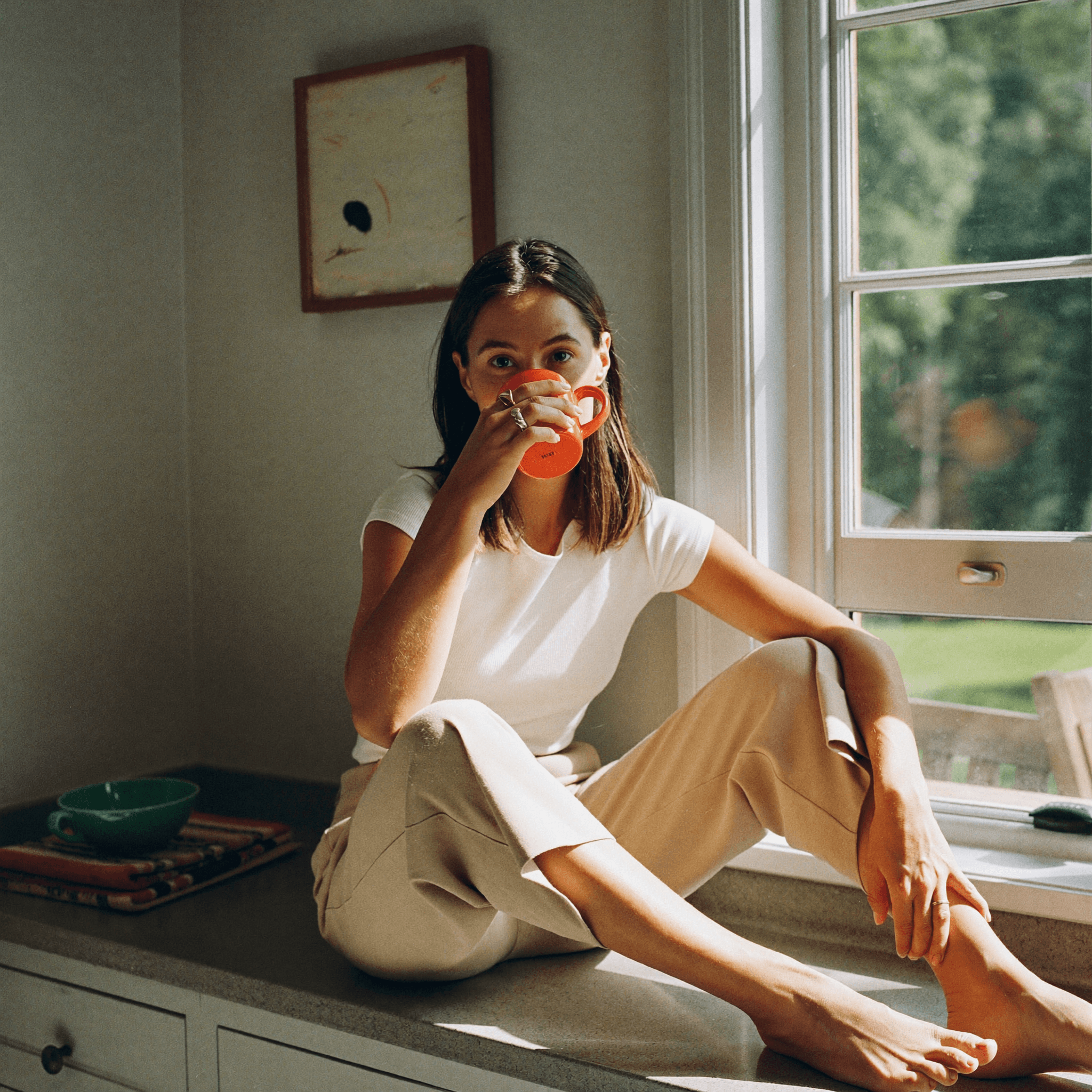 A woman sits on a kitchen window seat in soft mid-morning daylight, holding a mug